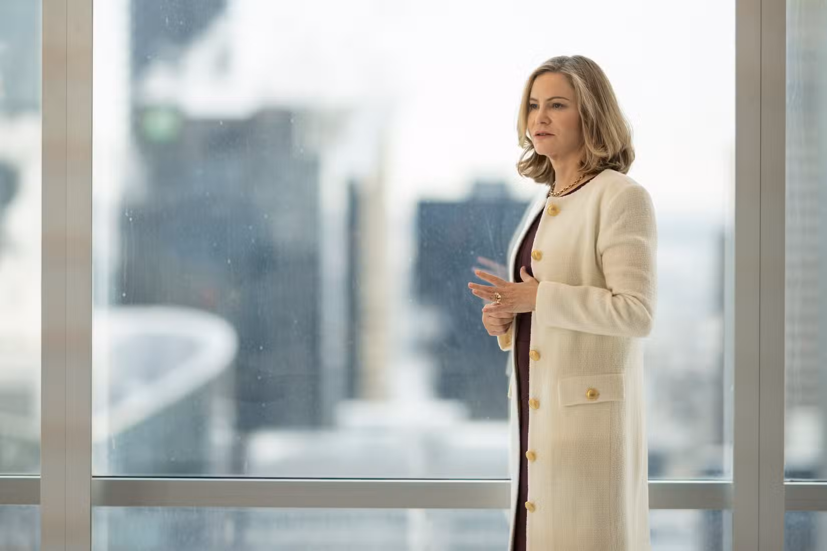 Jennifer Jason Leigh como Lorraine Lyon está com um casaco branco caro em frente à janela de um escritório na 5ª temporada de Fargo, da FX.