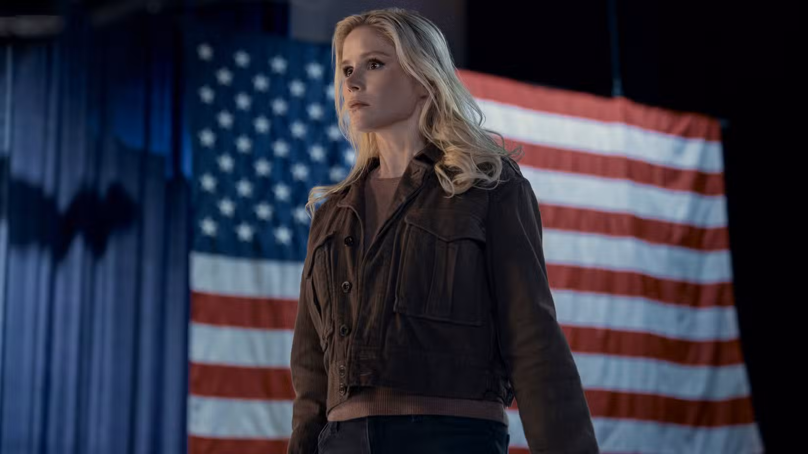 Annie January (Erin Moriarty) vestindo uma jaqueta marrom e jeans fica em frente a uma bandeira dos EUA na 5ª temporada de The Boys.
