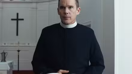 Um homem em traje de padre (Ethan Hawke) está atrás de um púlpito com uma cruz preta visível ao fundo.