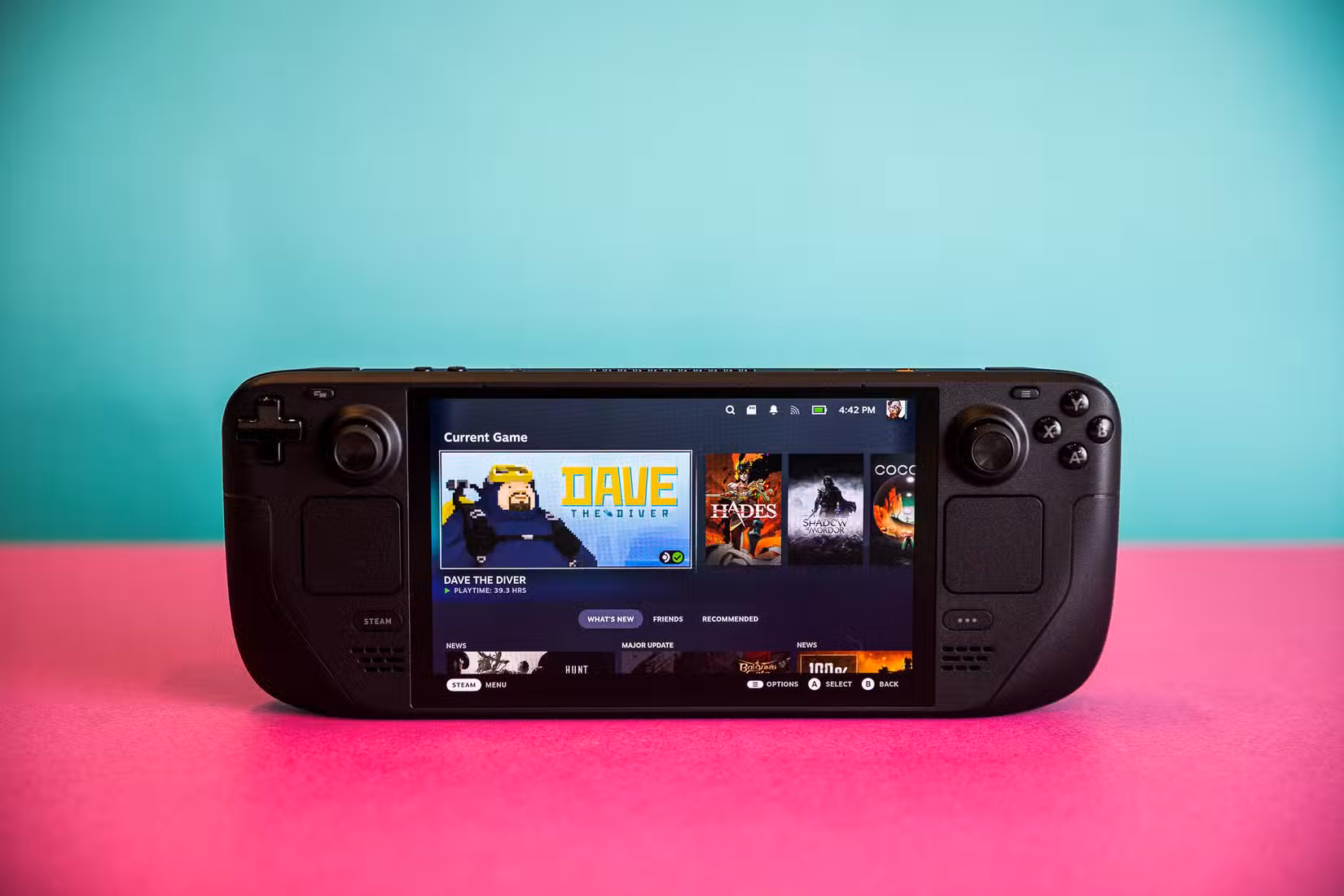 Fotografias do Steam Deck OLED e de seu case rígido com zíper. Cada foto é tirada com o computador de mão sobre uma folha de papel grosso rosa choque.