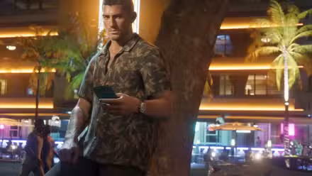 Um dos dois protagonistas de GTA 6, Jason, segura um telefone encostado em uma árvore. Ele está vestindo uma camisa estampada com folhas enquanto a vida noturna da cidade agita ao fundo. 