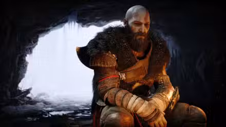 Kratos, vestindo pesadas peles de animais e com uma carranca no rosto, está sentado em frente a uma fogueira dentro de uma caverna. Atrás dele, podemos ver um mundo frio e invernal na entrada da caverna. 