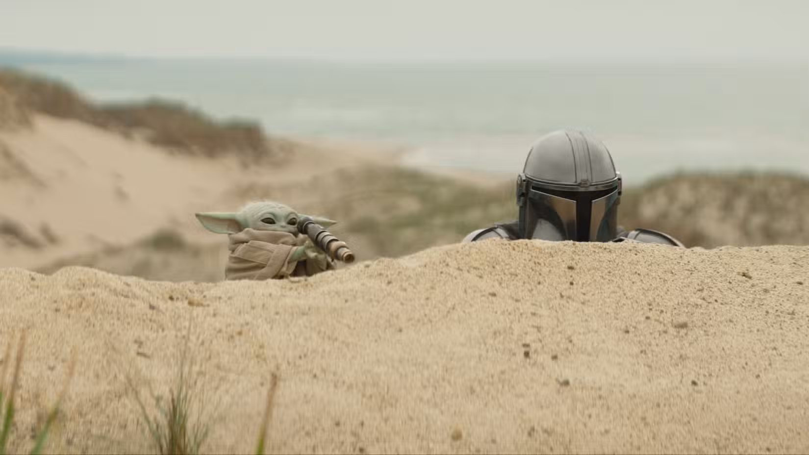 Grogu segura uma luneta enquanto espia uma duna de areia ao lado de Din Djarin em The Mandalorian e Grogu.