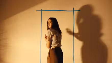 Mary (Renate Reinsve), uma mulher magra e pálida com longos cabelos pretos, fica de costas para a câmera, olhando nervosamente para trás por cima do ombro, em frente a um contorno de uma porta em uma parede em branco em Backrooms.