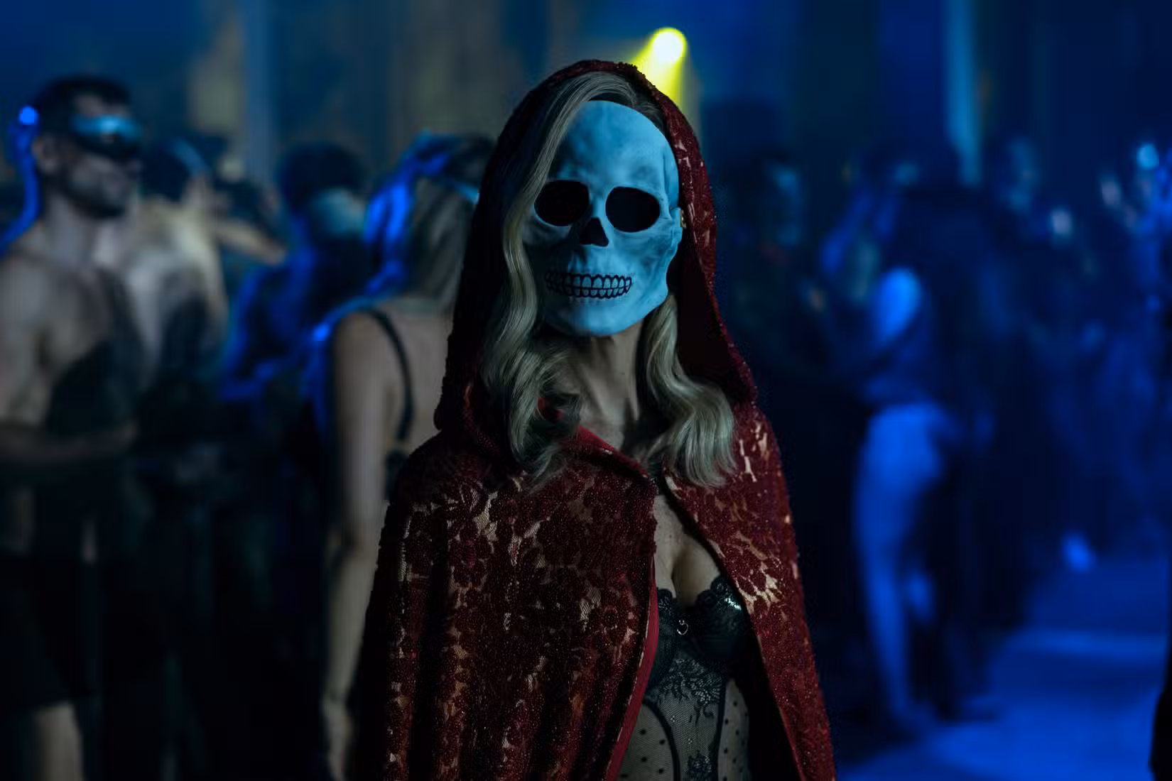 Carla Gugino como Verna, vestida com uma capa vermelha, vestido bondice de renda preta e uma máscara de caveira em A Queda da Casa de Usher.