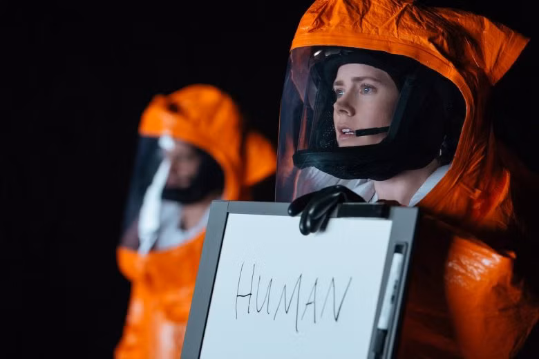 Amy Adams vestindo um traje anti-risco laranja e segurando um quadro branco com a palavra “humano” escrita em Chegada
