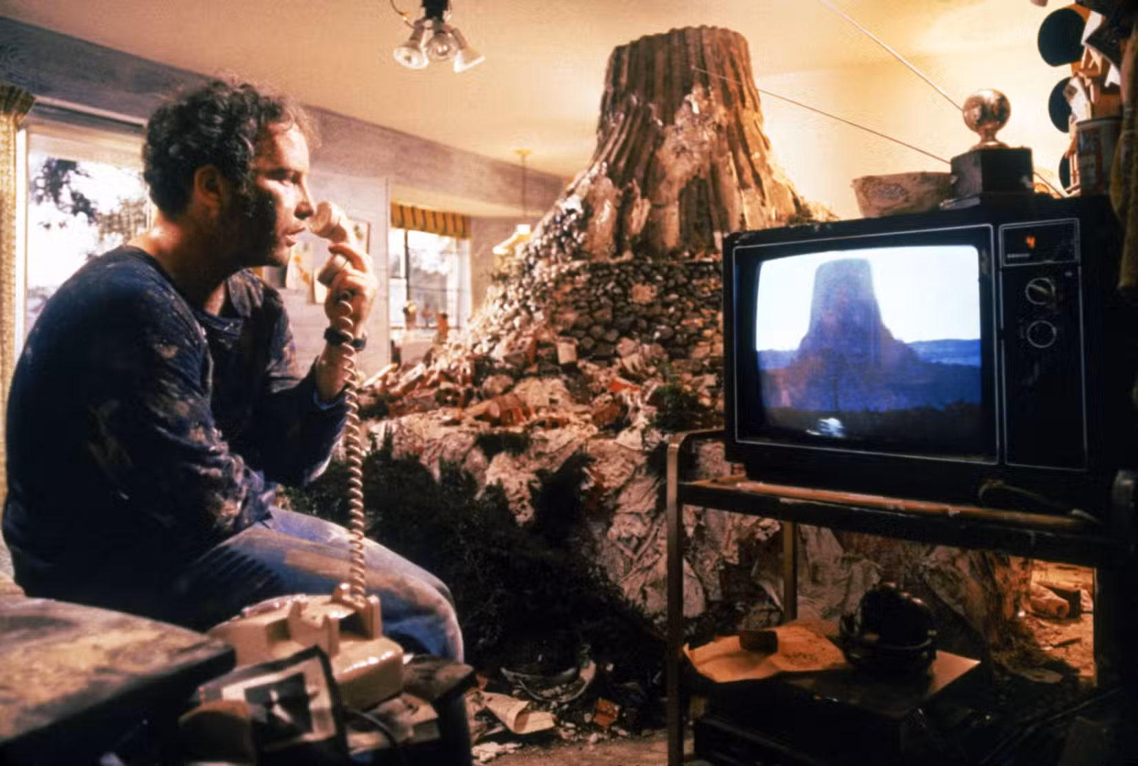 Richard Dreyfuss olha para a imagem de uma montanha em sua TV, com um enorme modelo de lama atrás dele