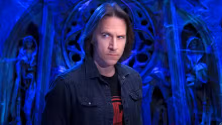 Uma imagem com Matthew Mercer da Critical Role. Ele está contra um fundo azul semelhante ao de uma igreja.