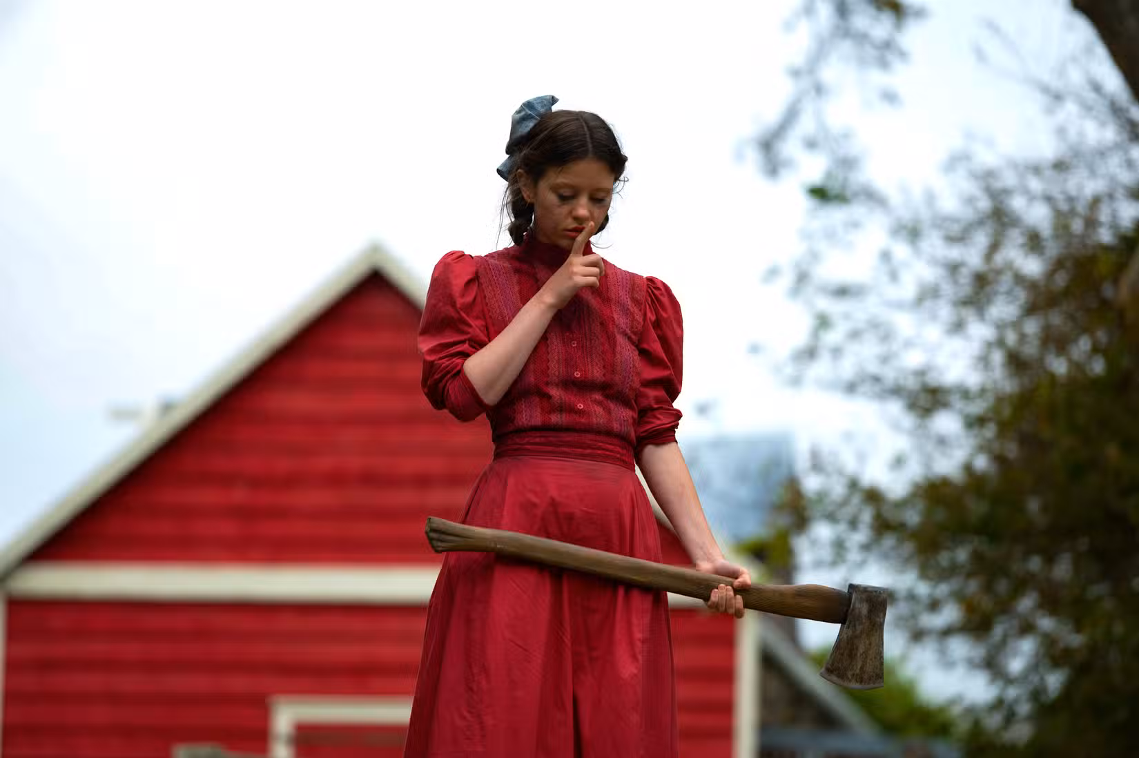 Mia Goth em um vestido vermelho vivo, segurando um machado e fazendo “Shush” com o dedo nos lábios em Pearl de Ti West