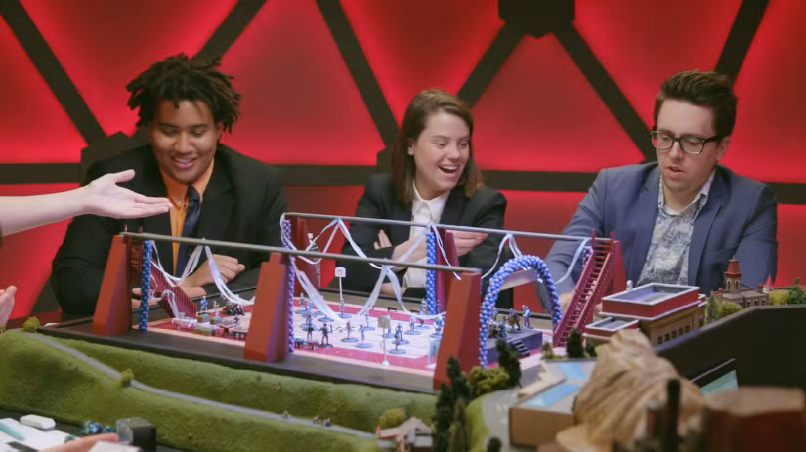 Lou Wilson, Ally Beardsley e Brian Murphy durante um episódio de Fantasy High. Há uma grande dança espalhada em miniatura na mesa.