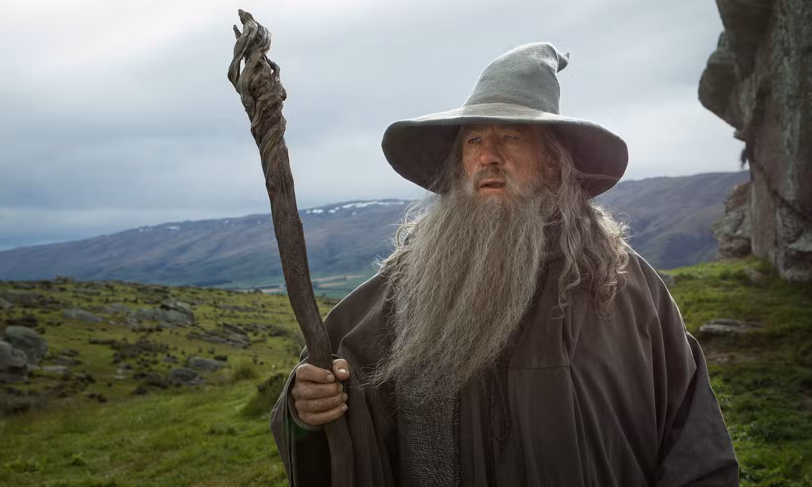 Gandalf, o Cinzento, segurando seu cajado com um campo verdejante e montanhoso atrás dele em O Hobbit: Uma Jornada Inesperada
