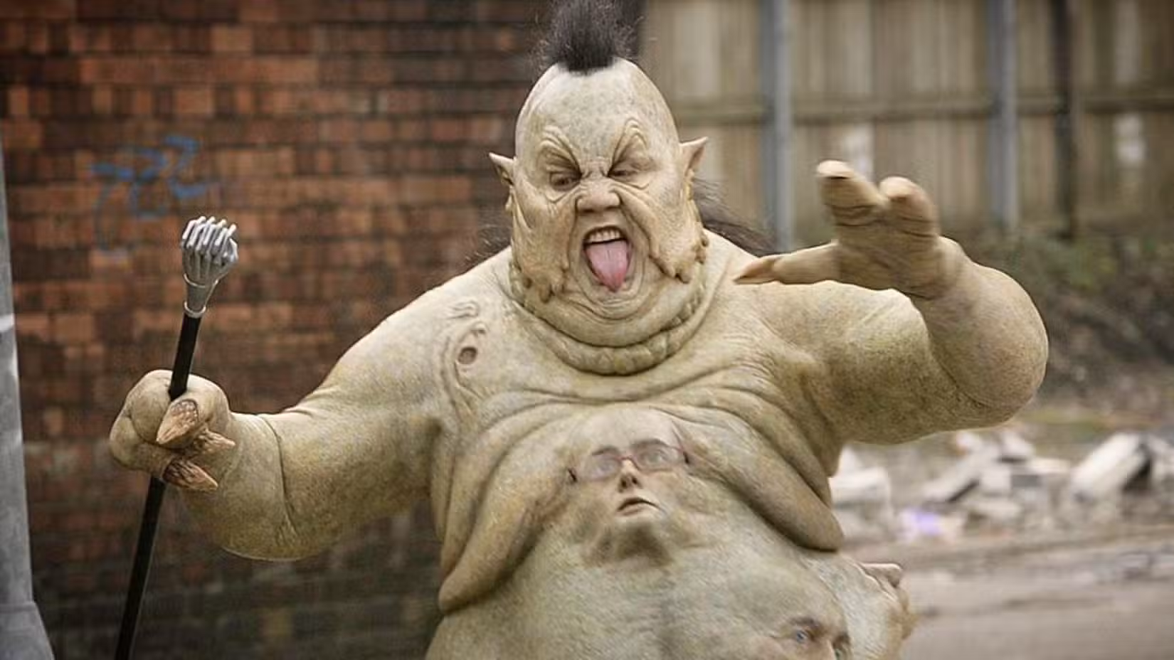 Uma imagem de Peter Kay como Abzorbaloff, um alienígena verde com uma bengala na mão. Ele corre para frente, a língua brandida para fora da boca.