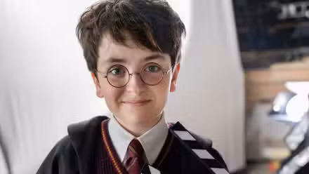 Dominic McLaughlin vestido como Harry Potter com suas vestes de Hogwarts, segurando uma tábua
