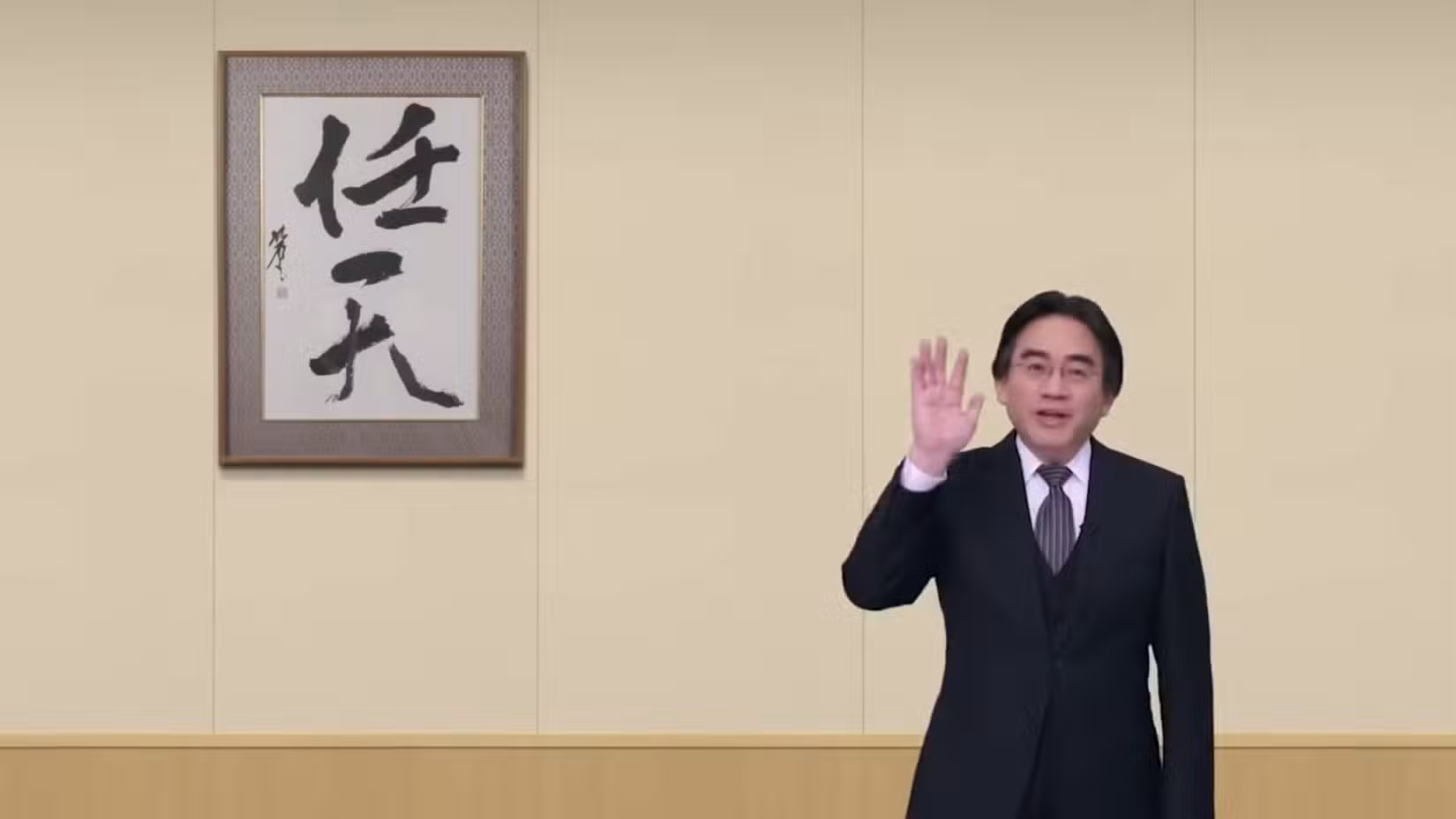 Satoru Iwata acena em frente a uma parede vazia com uma caligrafia japonesa emoldurada pendurada nela