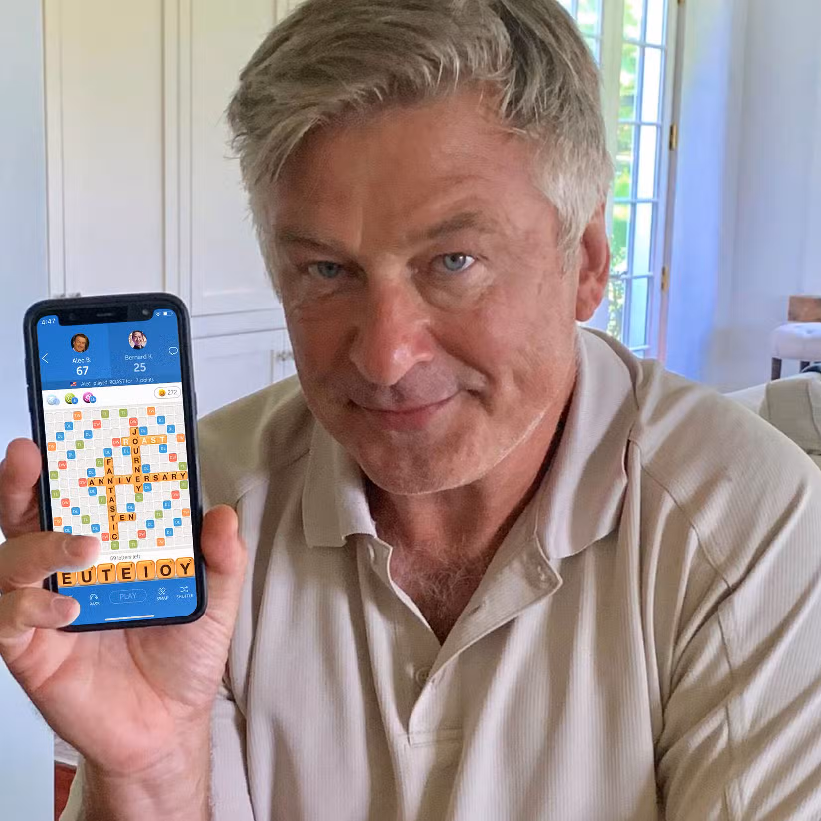 Um homem idoso - Alec Baldwin - com cabelos grisalhos e uma camisa pólo bege tem uma expressão predominantemente neutra no rosto. Ele está sorrindo levemente e segurando um iPhone com um jogo de Palavras com Amigos na tela.
