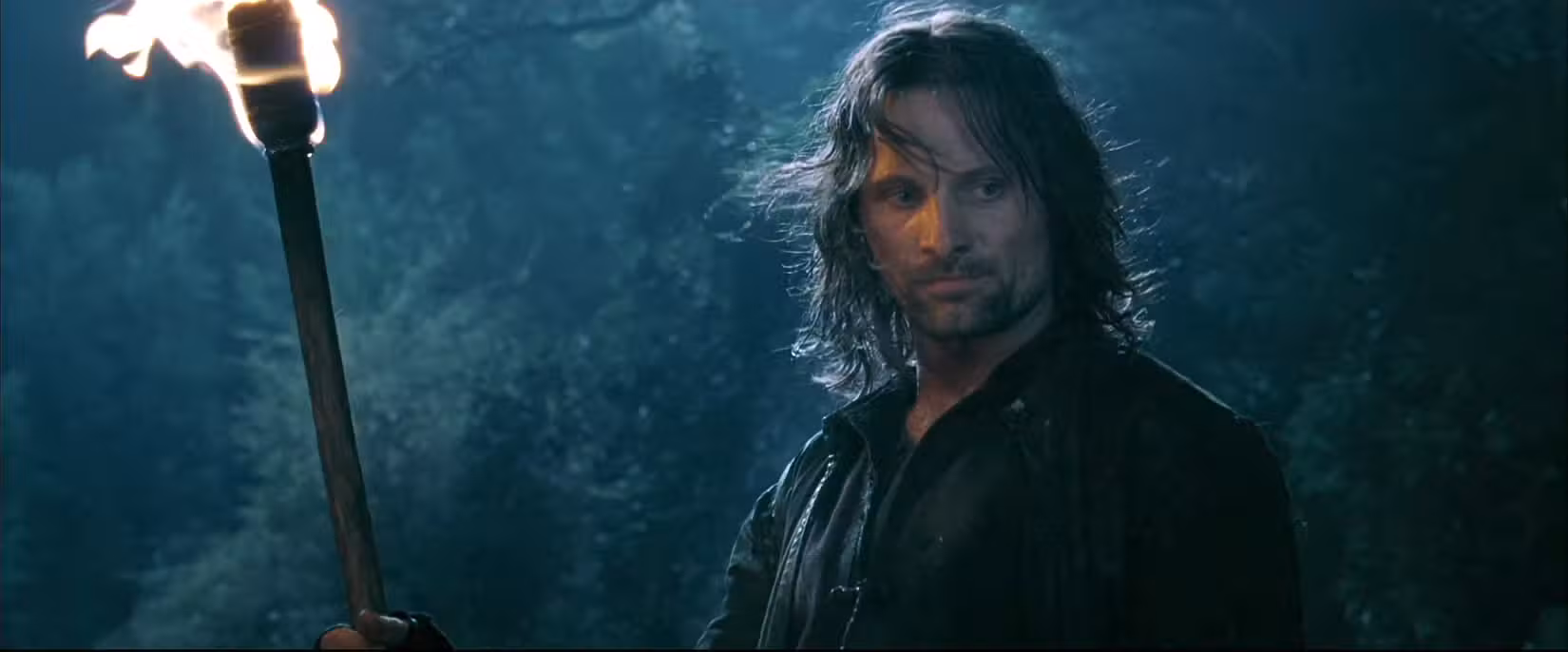 Um Aragorn enlameado está em uma floresta, segurando uma tocha acesa em A Sociedade do Anel.