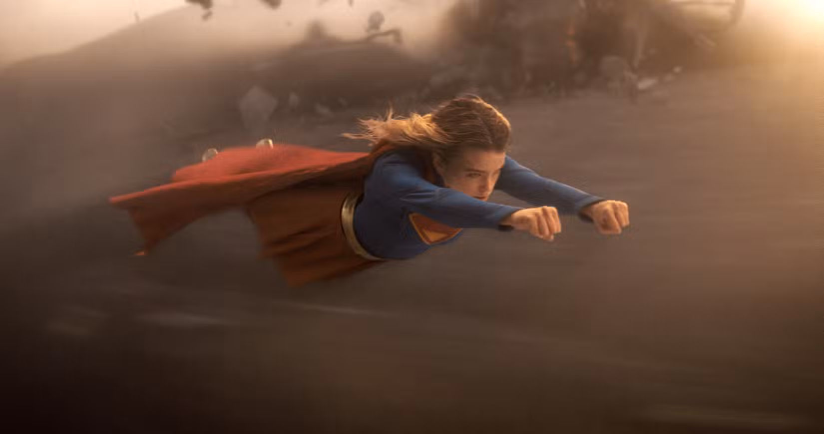 Milly Alcock como Supergirl, voando por uma paisagem arenosa, com os punhos cerrados à sua frente