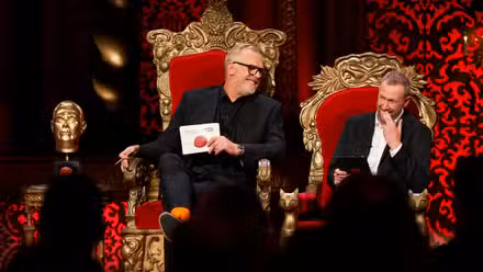 Greg Davies e Alex Horne riem em suas cadeiras de trono de tamanho cômico ao lado de uma estátua dourada da cabeça de Davies no set de Taskmaster.