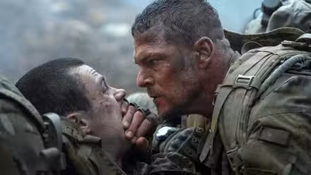 O sargento 81 do exército (Alan Ritchson) se inclina sobre um colega soldado e cobre sua boca com a mão para mantê-lo quieto em War Machine