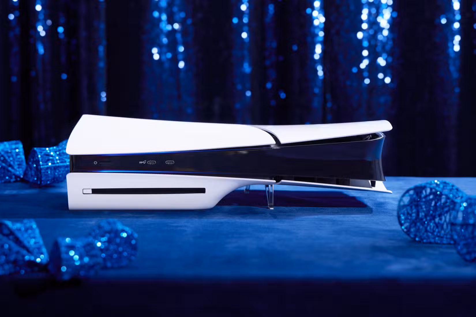 Uma fotografia do fino console PS5 da Sony colocado horizontalmente sobre uma superfície de camurça azul. Há enfeites festivos azuis ao redor do console que estão ligeiramente fora de foco, bem como uma cortina azul brilhante atrás do console.