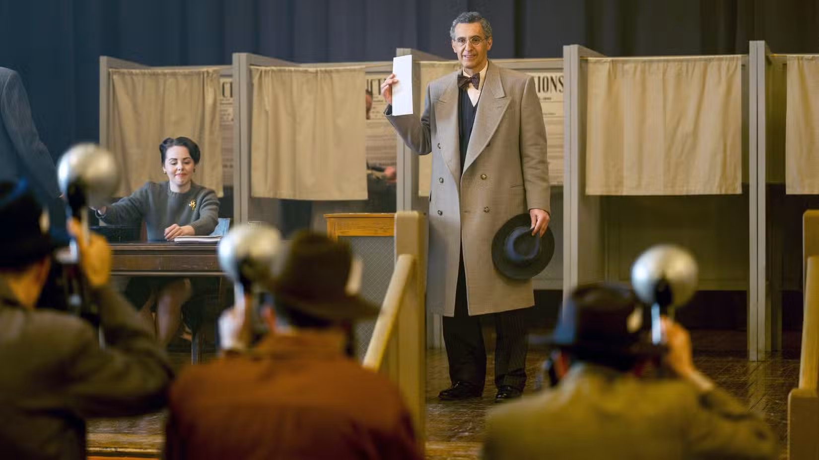 John Turturro realiza uma votação fora de uma cabine eleitoral enquanto repórteres tiram fotos no Plot Against America.