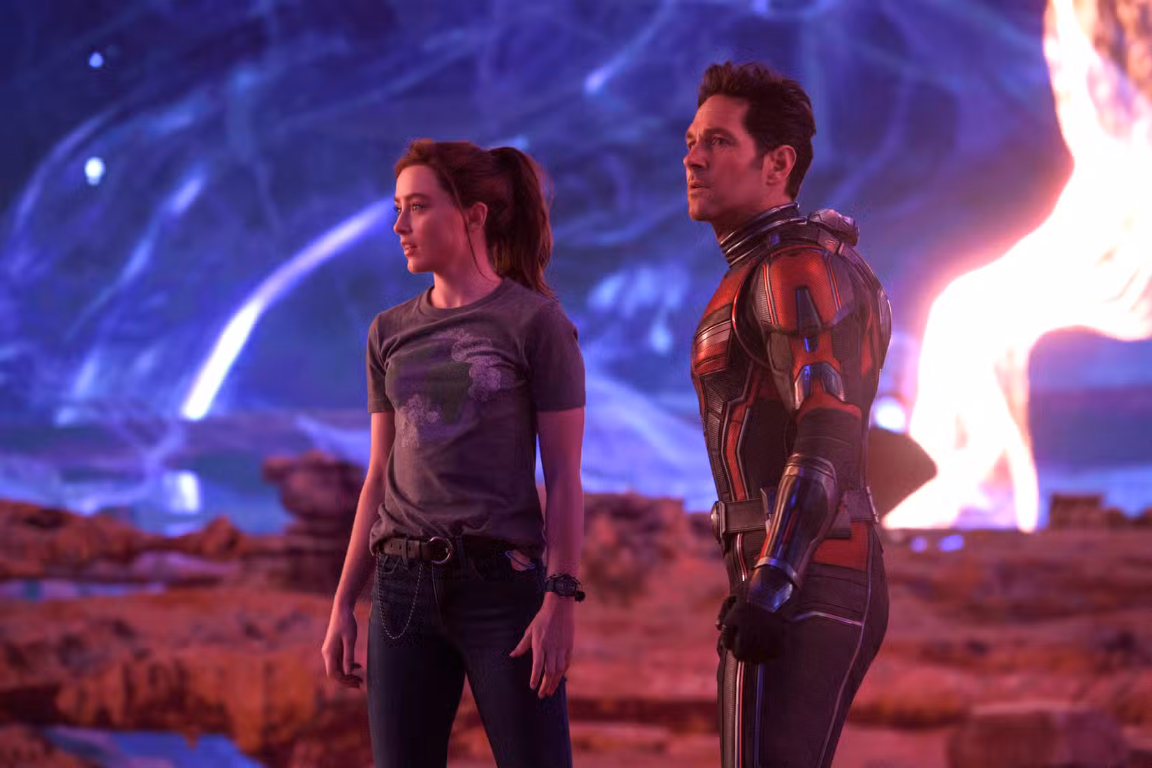 (LR): Kathryn Newton como Cassandra “Cassie” Lang e Paul Rudd como Scott Lang/Homem-Formiga em Homem-Formiga e a Vespa: Quantumania. Cassie está de jeans e camiseta, enquanto Scott está vestindo sua fantasia de Homem-Formiga sem capacete. Eles ficam atordoados em uma paisagem alienígena com céus rodopiantes.