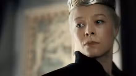 Uma imagem de House of the Dragon (HOTD) apresentando Emma D'arcy como Rhaenyra Targaryen. Ela usa uma coroa e olha para frente com severidade.
