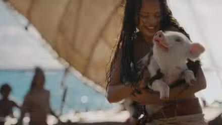 Moana (Catherine Laga'aia), uma adolescente das ilhas do Pacífico, está na frente de um pequeno navio segurando seu porco de estimação Pua no live-action da Disney Moana