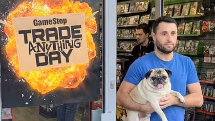 um homem carrega um pug ao lado de um anúncio de comércio de qualquer coisa do GameSpot todos os dias