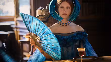 Mia Goth usando um vestido azul e segurando um leque combinando em Frankenstein