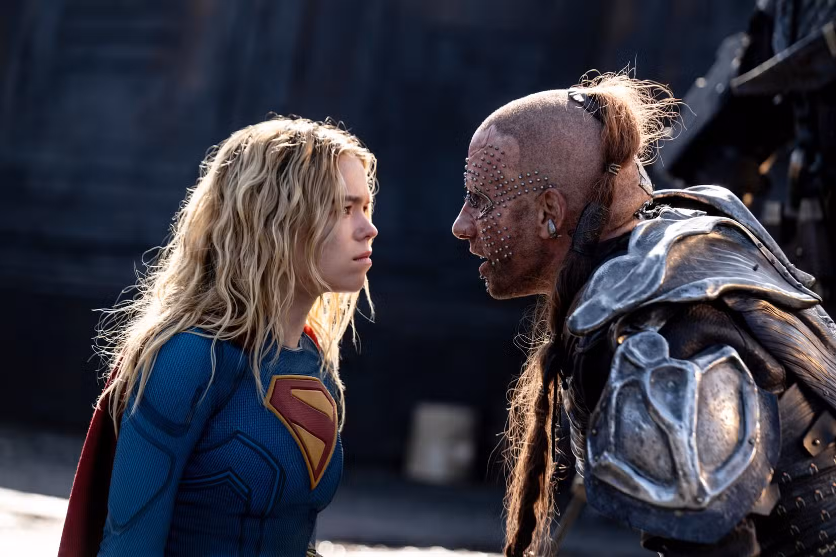 Milly Alcock como Supergirl, no traje tradicional, enfrenta Krem, um homem vestindo uma armadura de metal remendada, cabeça raspada e longo rabo de cavalo com topete, e uma extensa grade de piercings faciais