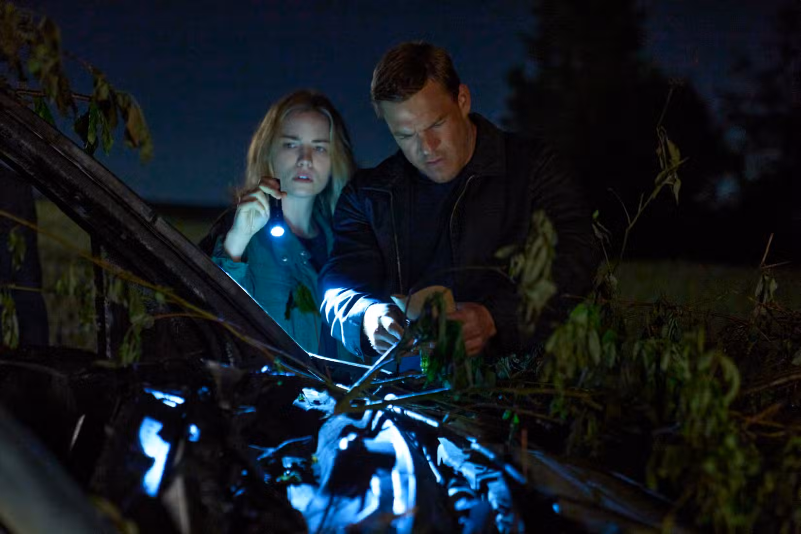 Jack Reacher, um homem grande vestindo um suéter de gola, investiga na floresta com uma policial loira (segurando uma lanterna).
