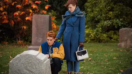 Archie (KJ Apa) ajoelhado em frente a um túmulo enquanto sua mãe coloca a mão em seu ombro
