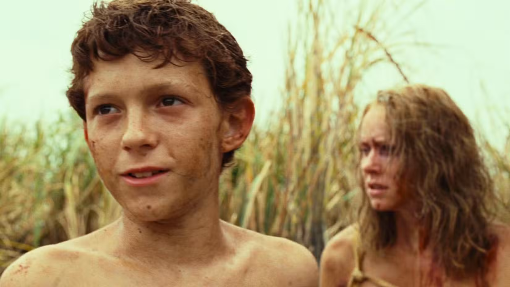 Tom Holland, sujo e sombrio, olha para frente enquanto Naomi Watts olha ao fundo.