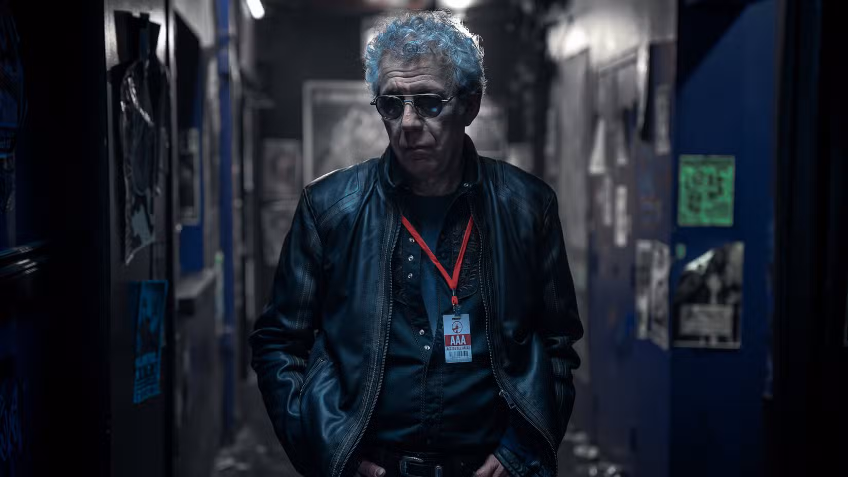 Daniel Molloy (Eric Bogosian) usa óculos escuros, uma jaqueta de couro e um distintivo em um cordão vermelho enquanto caminha nos bastidores de O Vampiro Lestat