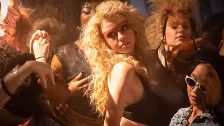 Maxine (Mia Goth), uma máscara retocada em seu rosto e seu cabelo penteado em uma grande nuvem loira, dança com um grupo de estranhos em uma boate em Ti West's Maxxine.