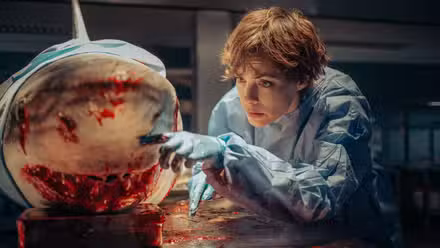 A pesquisadora oceânica Sophia (Bérénice Bejo) se inclina para observar de perto o focinho sangrento de um grande tubarão que ela está dissecando em um laboratório no thriller de tubarão da Netflix de Xavier Gens, Under Paris